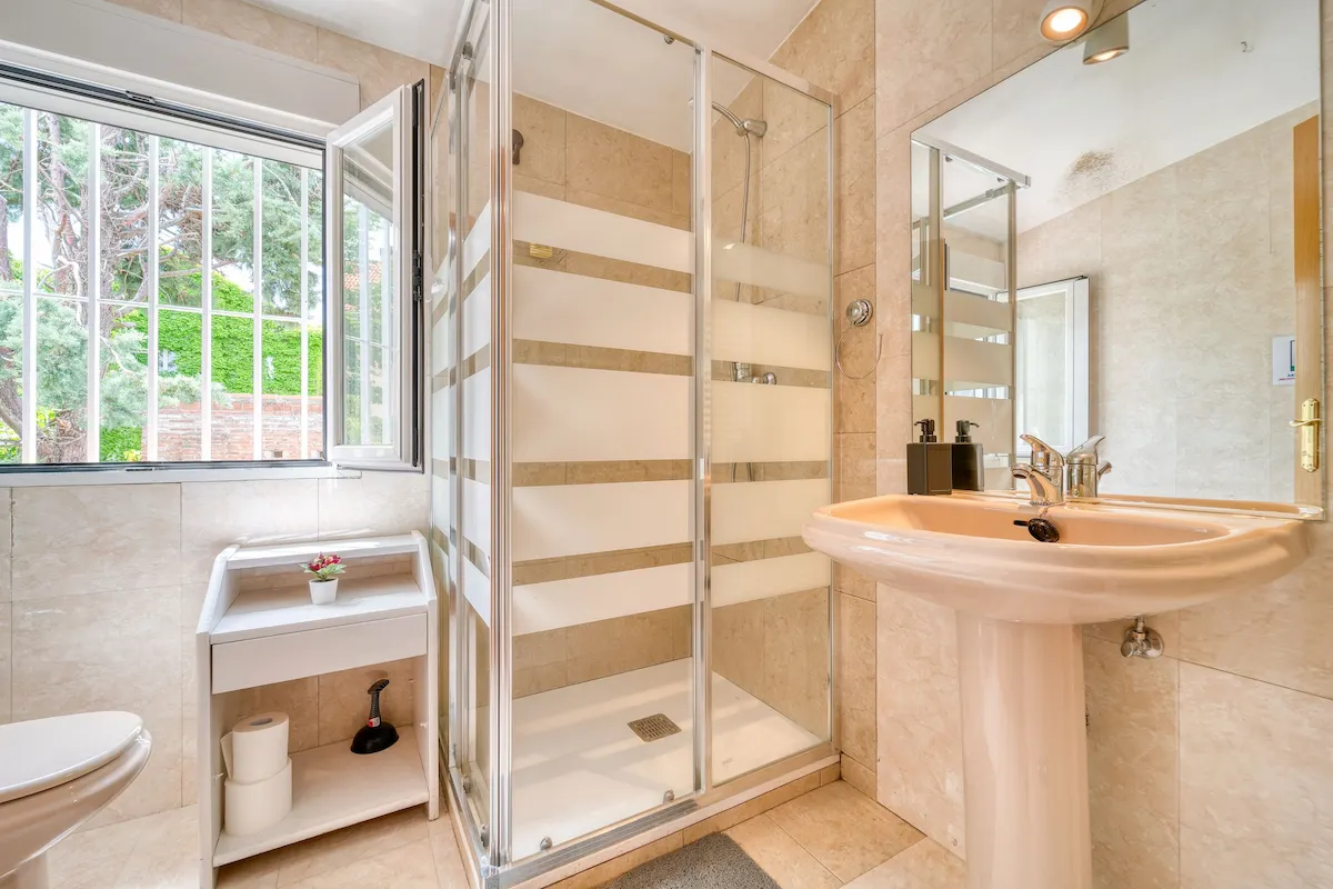 Bright beige bathroom with a glass shower, pedestal sink, and an open window overlooking greenery.