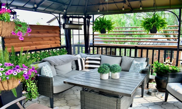 Covered outdoor patio with gray seating, hanging ferns, flowering plants, and a large coffee table.