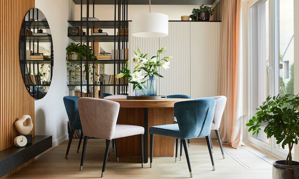 Modern dining room with a wooden table, blue and beige chairs, white lilies, open shelving, and soft natural light.